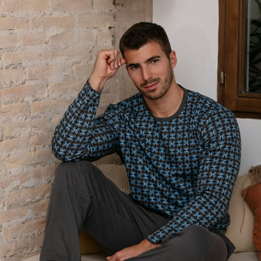 Man sitting on a couch wearing a blue patterned shirt and gray pants, with a plant and brick wall in the background.